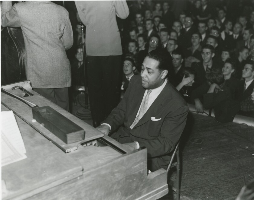 In a black and white photo, Duke Ellington plays a piano in front of a crowded room.